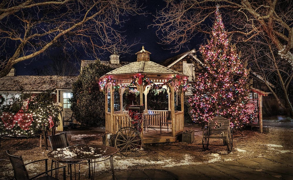 A gazebo in a backyard decorated with multicolor Christmas lights on the trees and bushes.