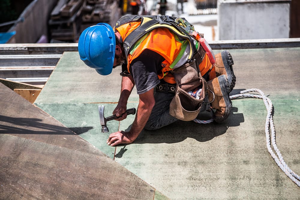 A man in a blue hard hat and orange vest on his knees hammering a nail into an unfinished roof.