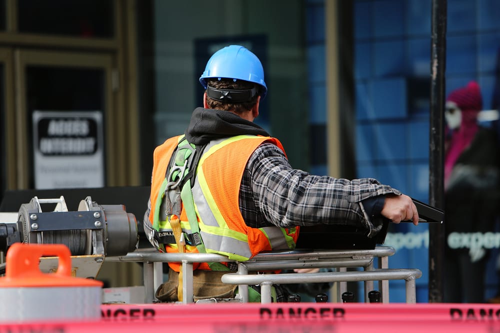 A construction worker wearing a blue hard hat and orange vest looking at a construction site. Red danger tape is in the foreground.