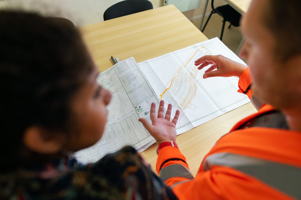 Two people discussing blueprints and data lying on a wooden table in front of them.