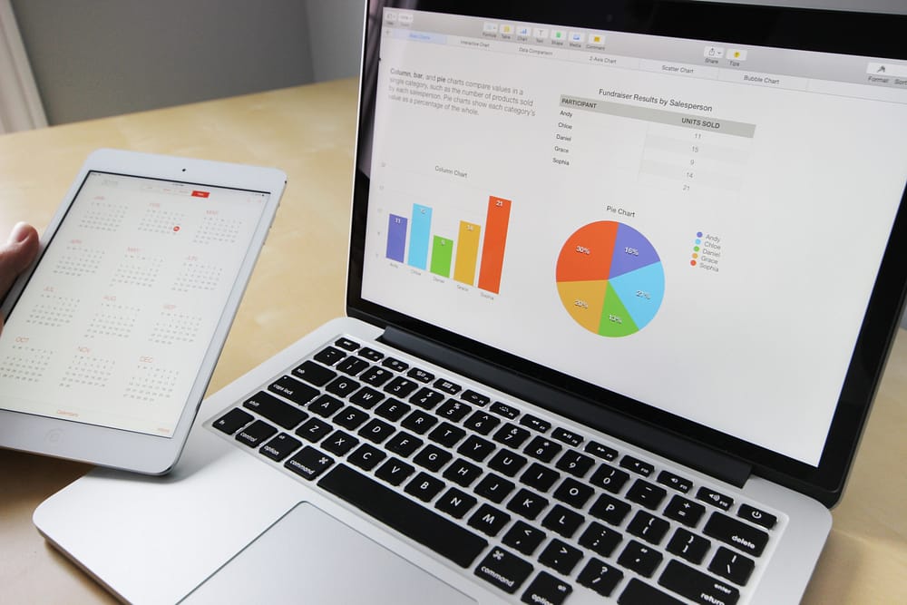 A laptop displaying multicolor graphs and someone holding a tablet to the left displaying a yearly calendar.