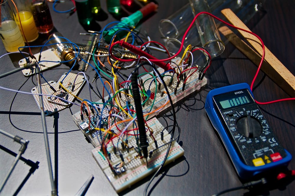 A black table with parts of an electrical board scattered across. Multicolored wires are tangled above the board.