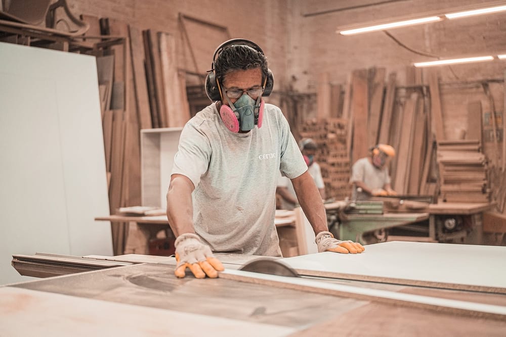 A person wearing a pink respirator sawing a large panel of wood in a warehouse.