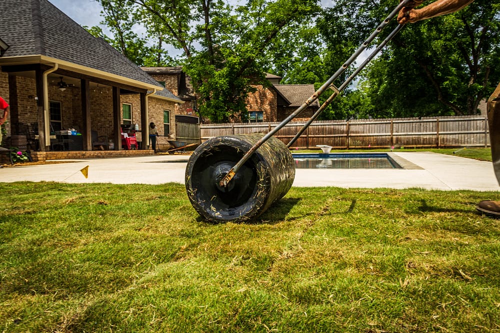 A person using a lawn roller to level their backyard with a swimming pool and wooden fence.