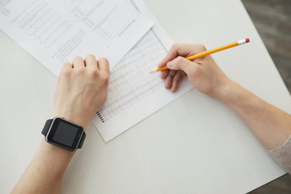 A person wearing a black smartwatch writing on a white piece of paper with a yellow pencil in their right hand.