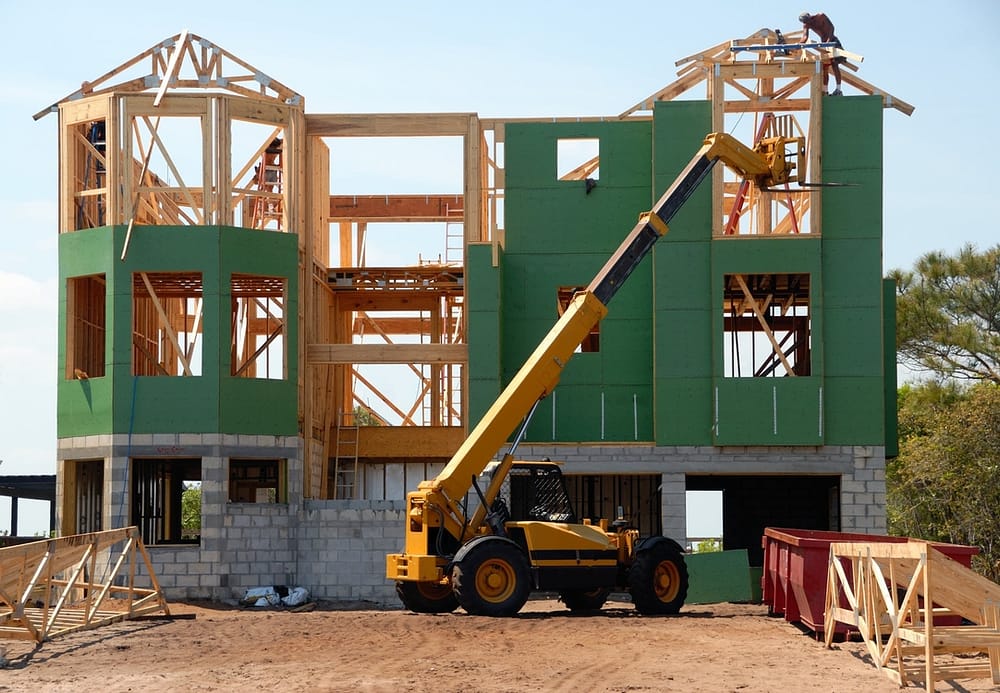 A green home under construction with a yellow lift sitting in the front. 