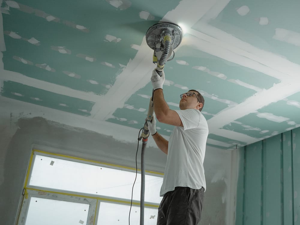 A man in a white t-shirt and gloves polishing a light blue ceiling with white markings.