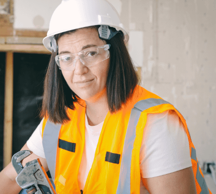 A person with shoulder-length brown hair wearing safety glasses, a white hard hat, and an orange safety vest. 