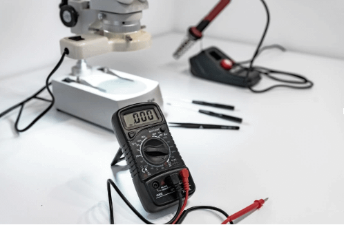 A black multimeter on a white tabletop with other electrical devices in the background.