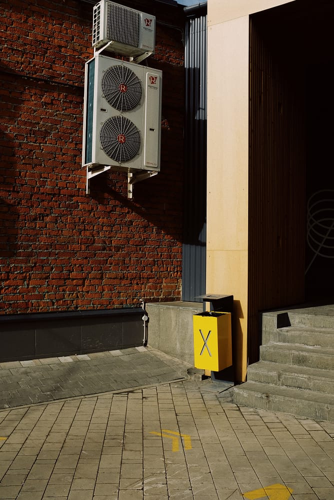 An HVAC plenum hanging on an red brick wall. A trash can with a yellow X sits underneath.