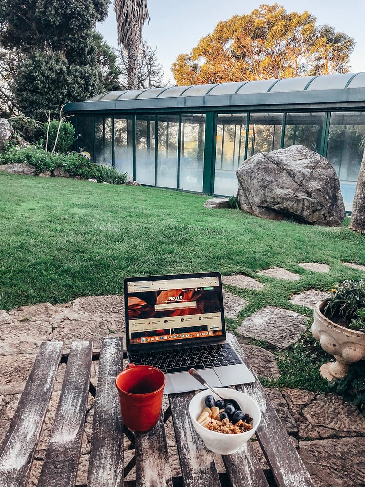 A wooden table with a computer, coffee mug, and bowl of granola with a stone sidewalk and greenhouse in the background.