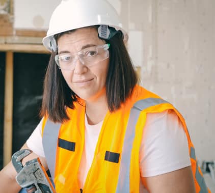 A person with shoulder-length brown hair wearing safety glasses, a white hard hat, and an orange safety vest.